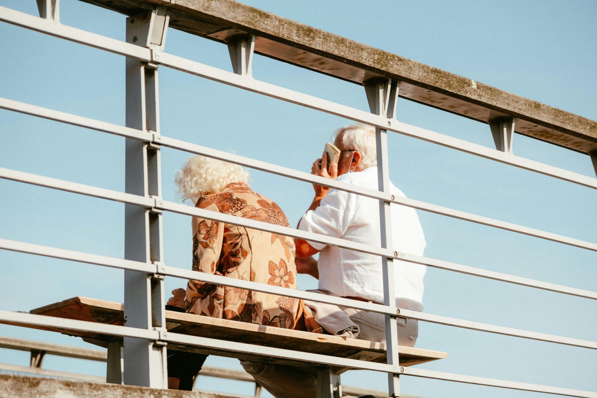 Two people sitting on a bridge, one in a white outfit and one in a patterned dress, against a blue sky