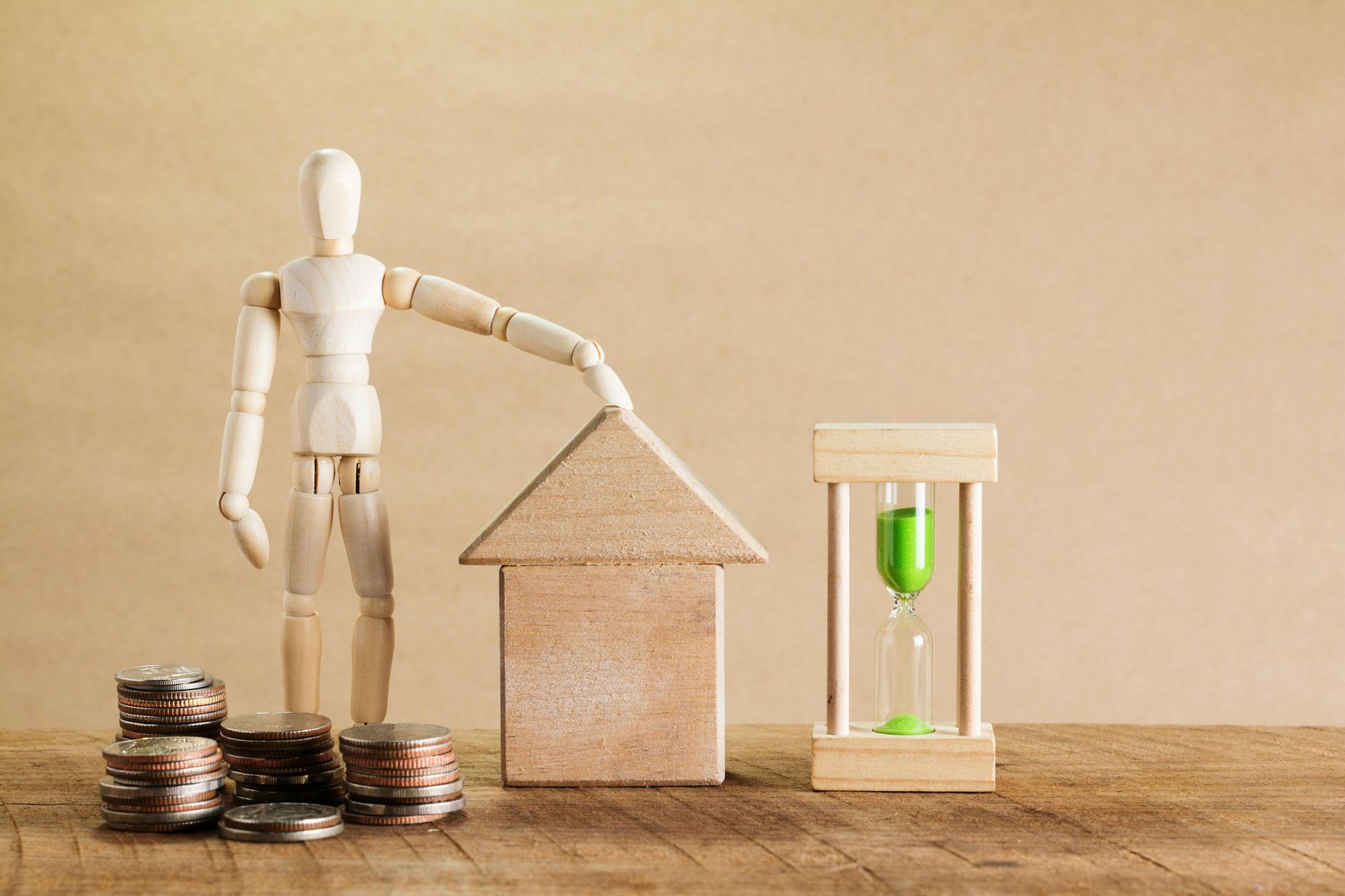 Wooden mannequin beside house model, coin stacks, and hourglass on a tabletop, symbolizing savings and time