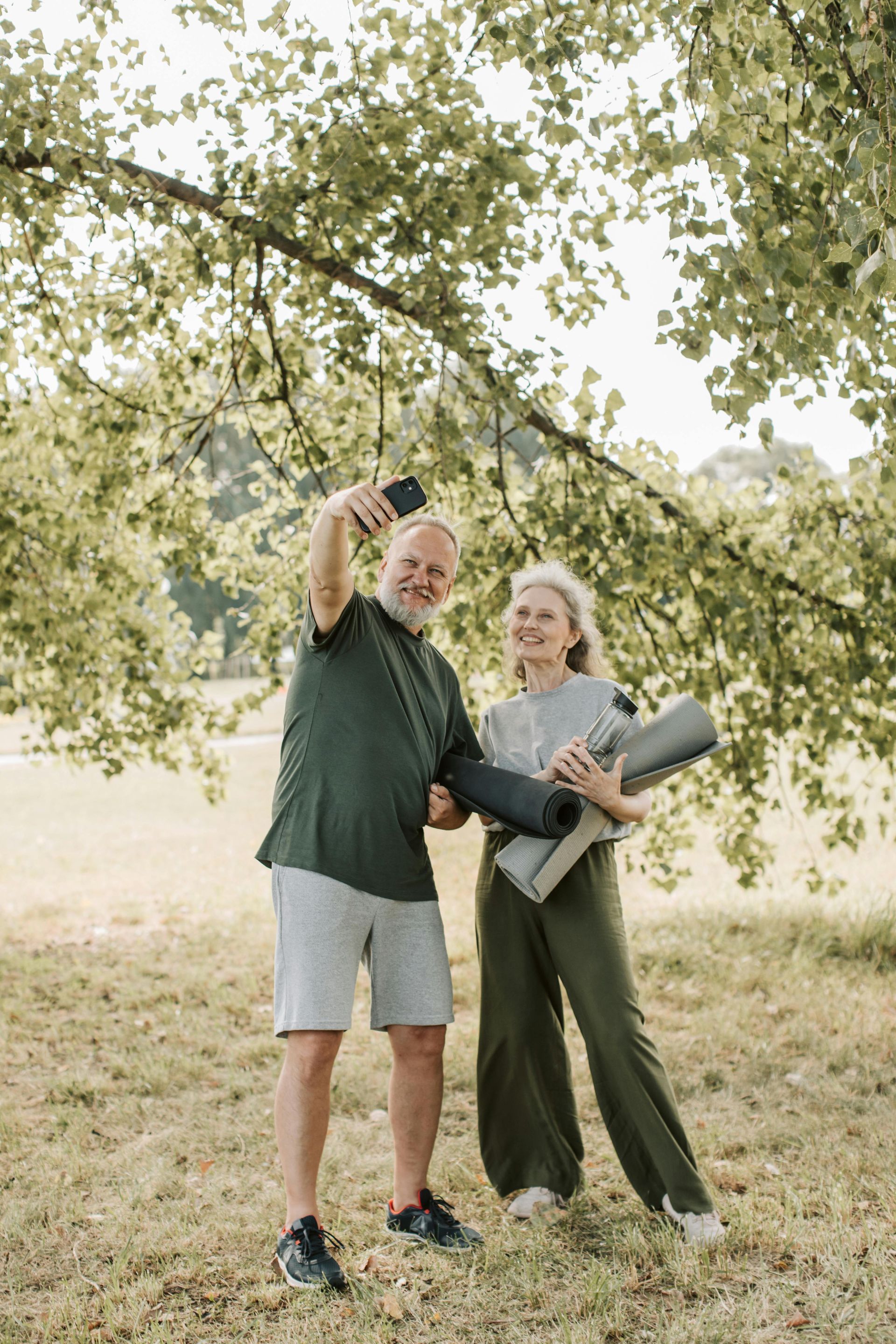 Two people stand under a tree, smiling and pointing upward, holding rolled yoga mats in a grassy field.