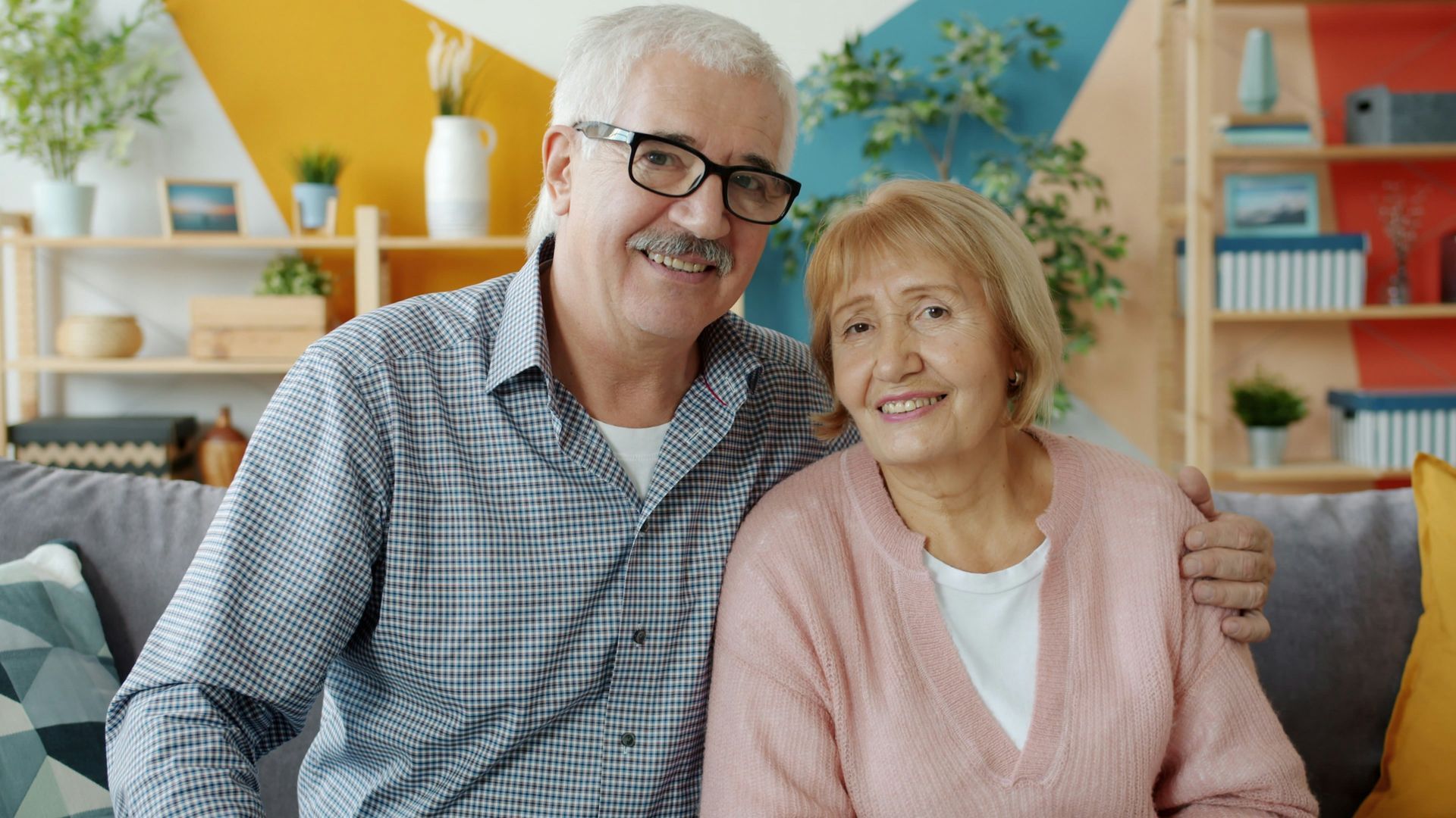Smiling couple sitting together on a couch in a bright living room with plants and shelves behind them