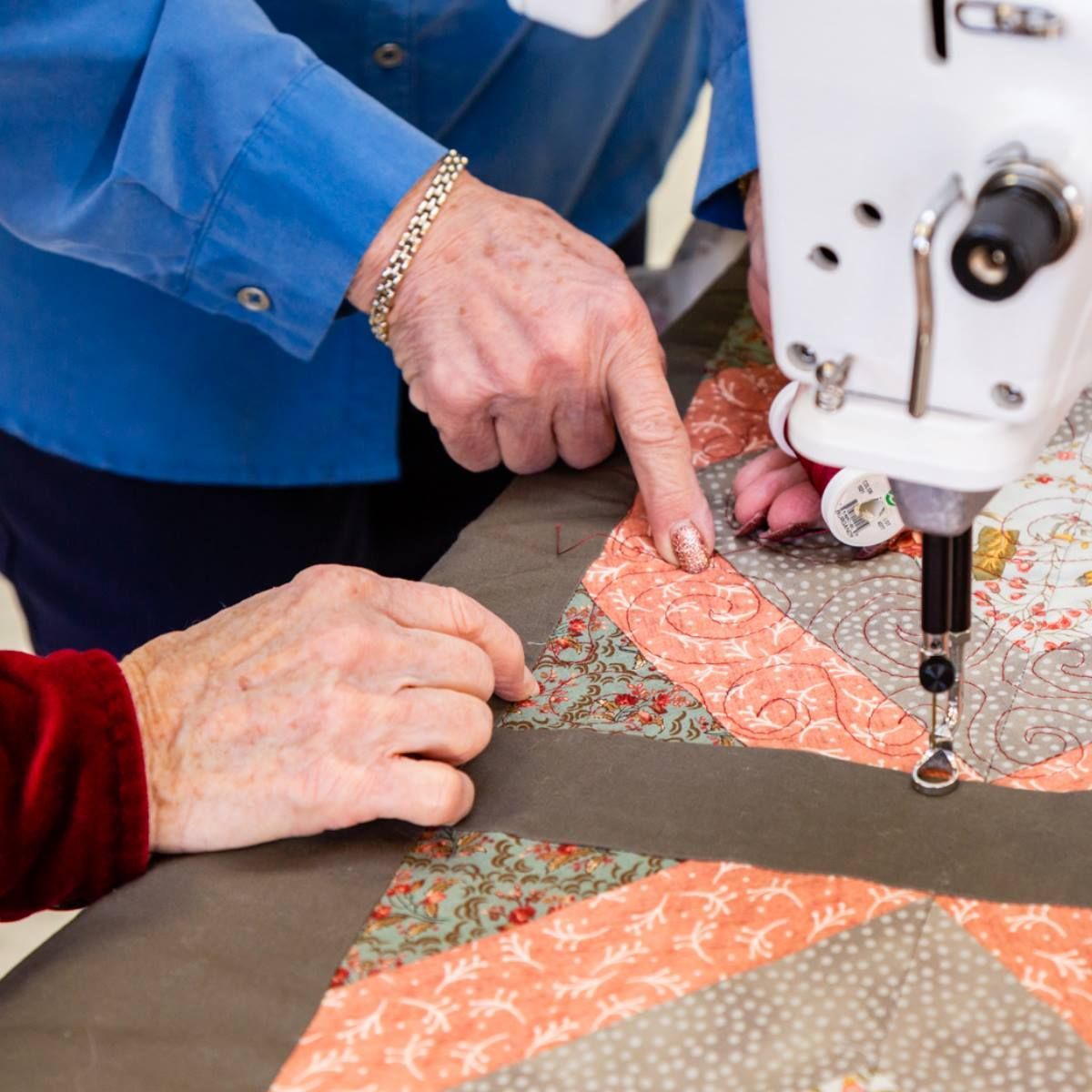 Hands guiding quilt fabric under a sewing machine as a patterned quilt is being stitched.