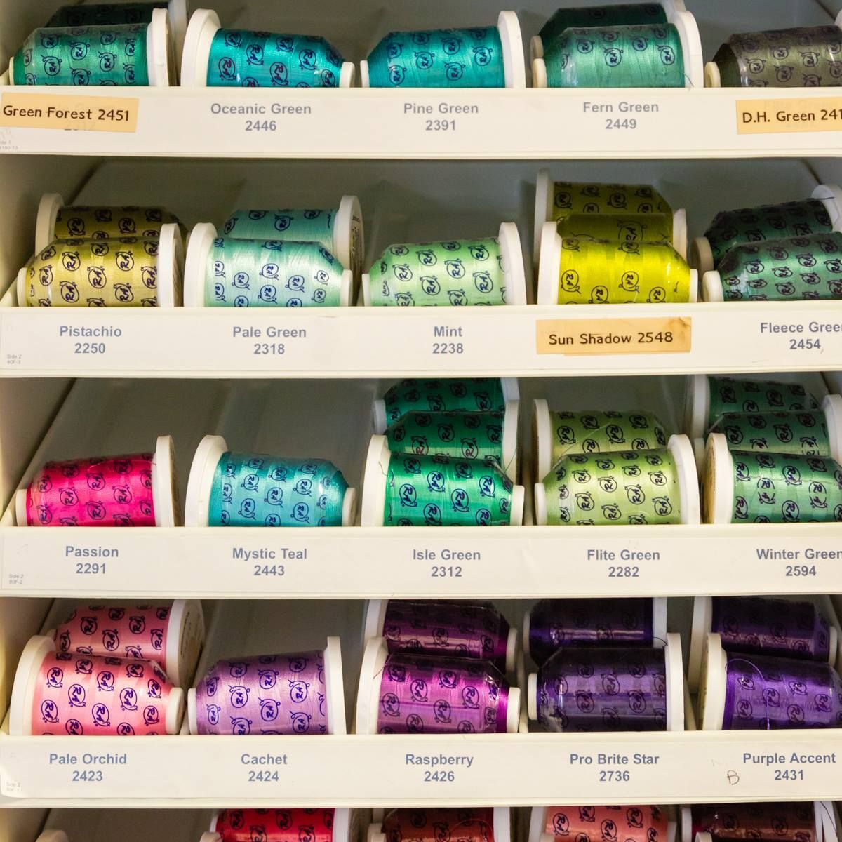 Rows of colorful thread spools on shelves, labeled by color name in a sewing shop.