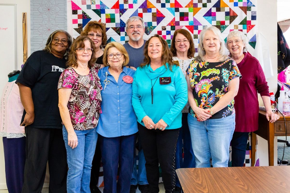Group of people standing indoors in front of a colorful quilt display, smiling at the camera.