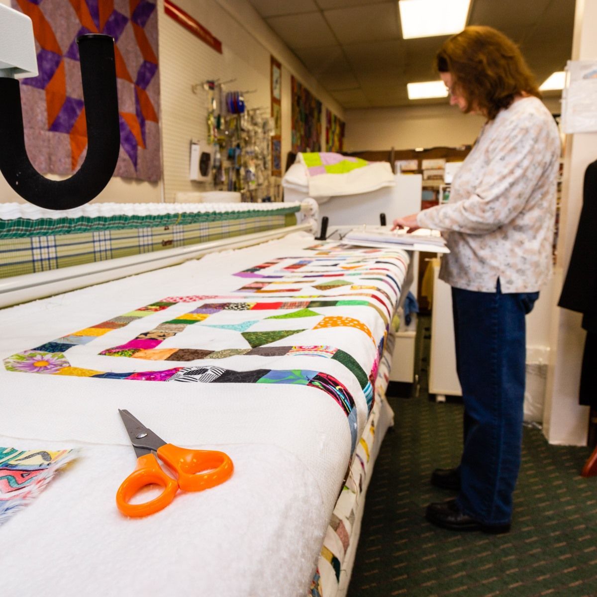 Quilter sewing a colorful patchwork quilt on a long machine, with orange scissors in the foreground.
