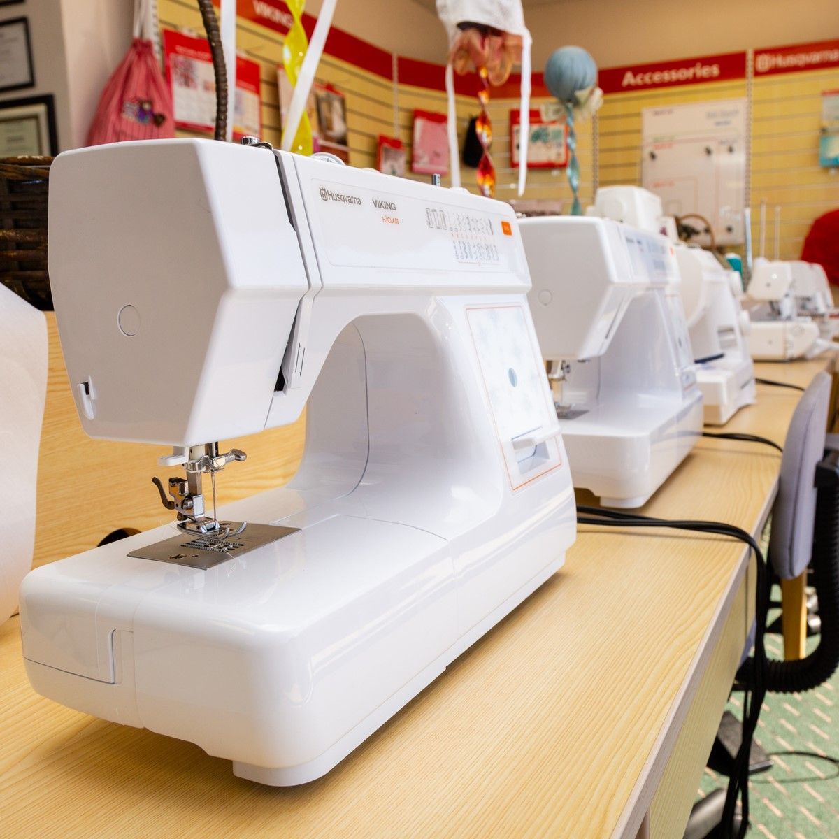 Row of white sewing machines on a store display table, with accessories on shelves in the background