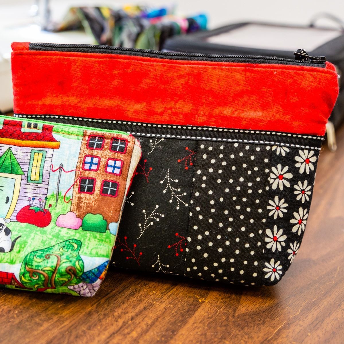 Two small fabric zip pouches on a wooden table, one red and one black with floral print, plus a colorful pouch in front