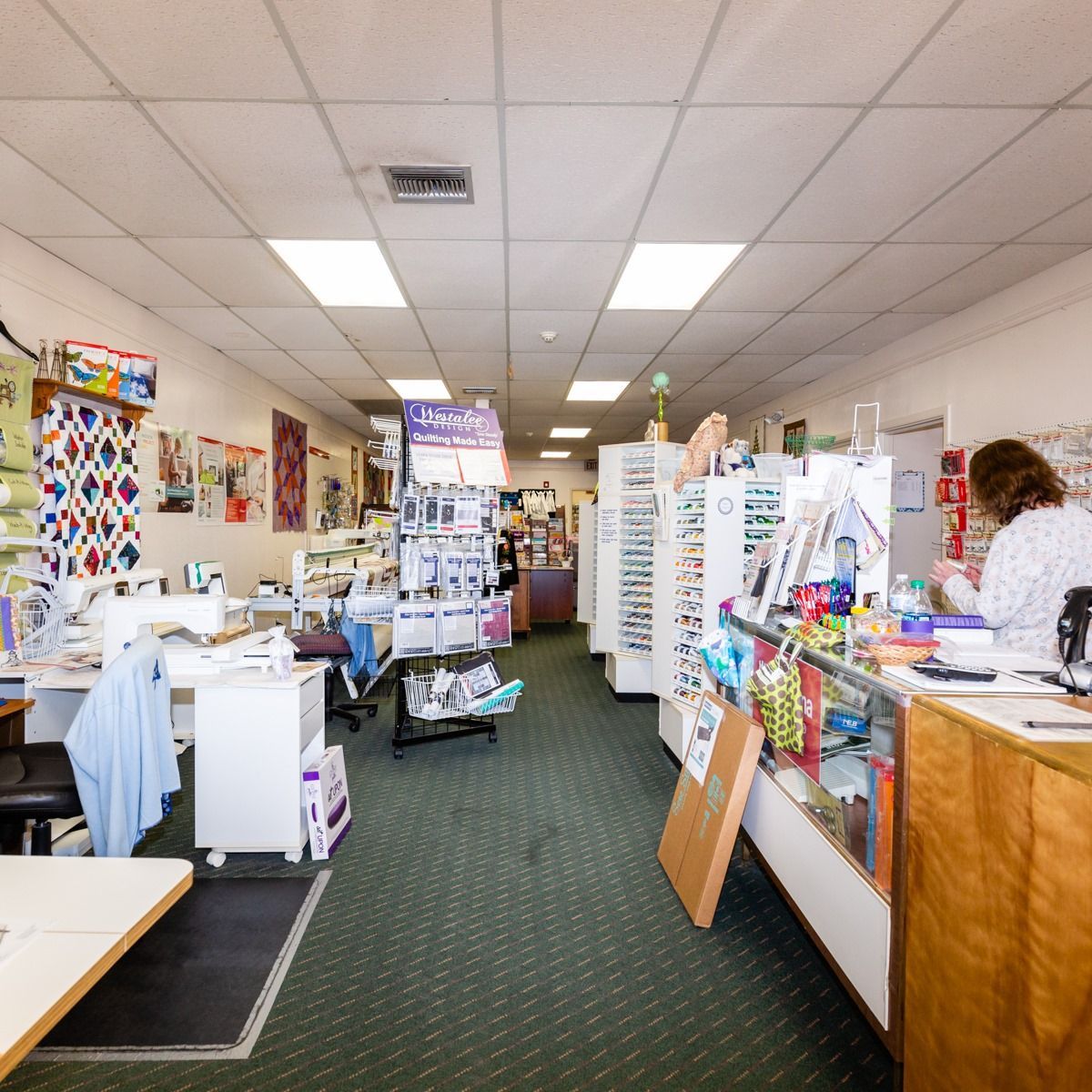 Bright retail shop interior with shelves of cards, gifts, and supplies; a person stands at the counter.
