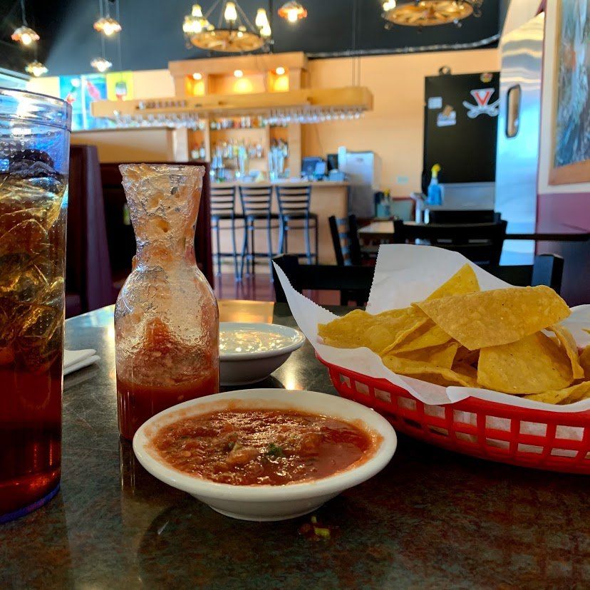 Table setting at a Mexican restaurant: salsa, chips, bottle, drink, and background bar.