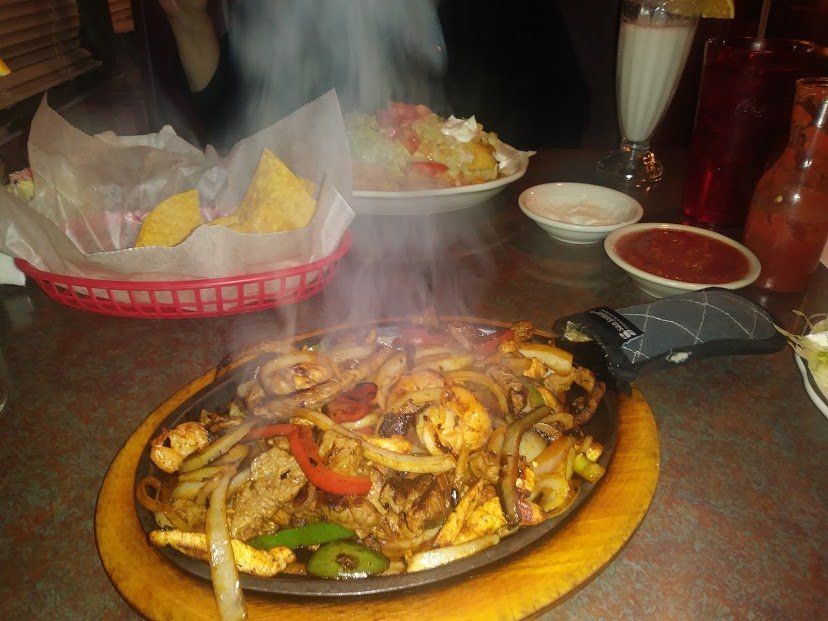 Sizzling fajitas on a wooden platter with other Mexican food, drinks, and a basket of chips on a table.
