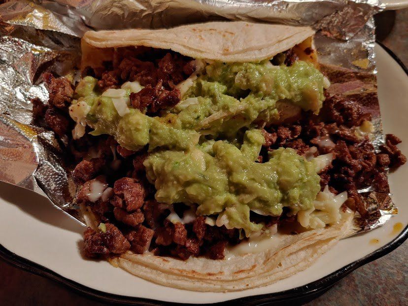 Taco with meat and guacamole on a tortilla, served on a plate lined with foil.