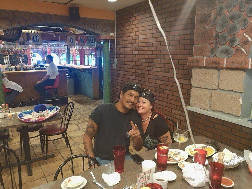 Couple at a Mexican restaurant smiles, table of food in front of them, server at bar in background.