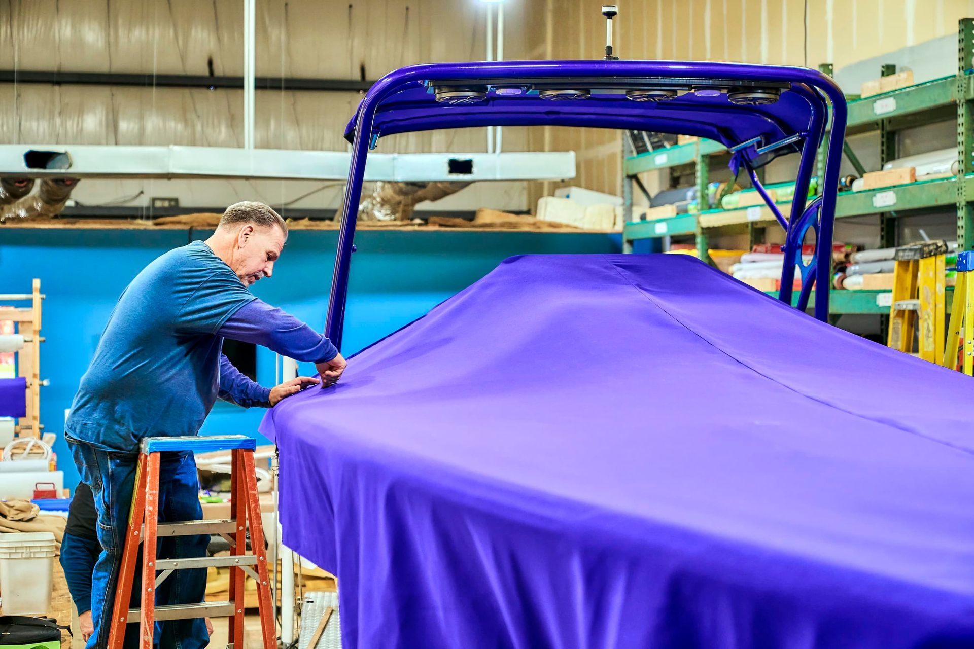 Man on ladder fitting purple fabric cover onto a boat in a workshop.