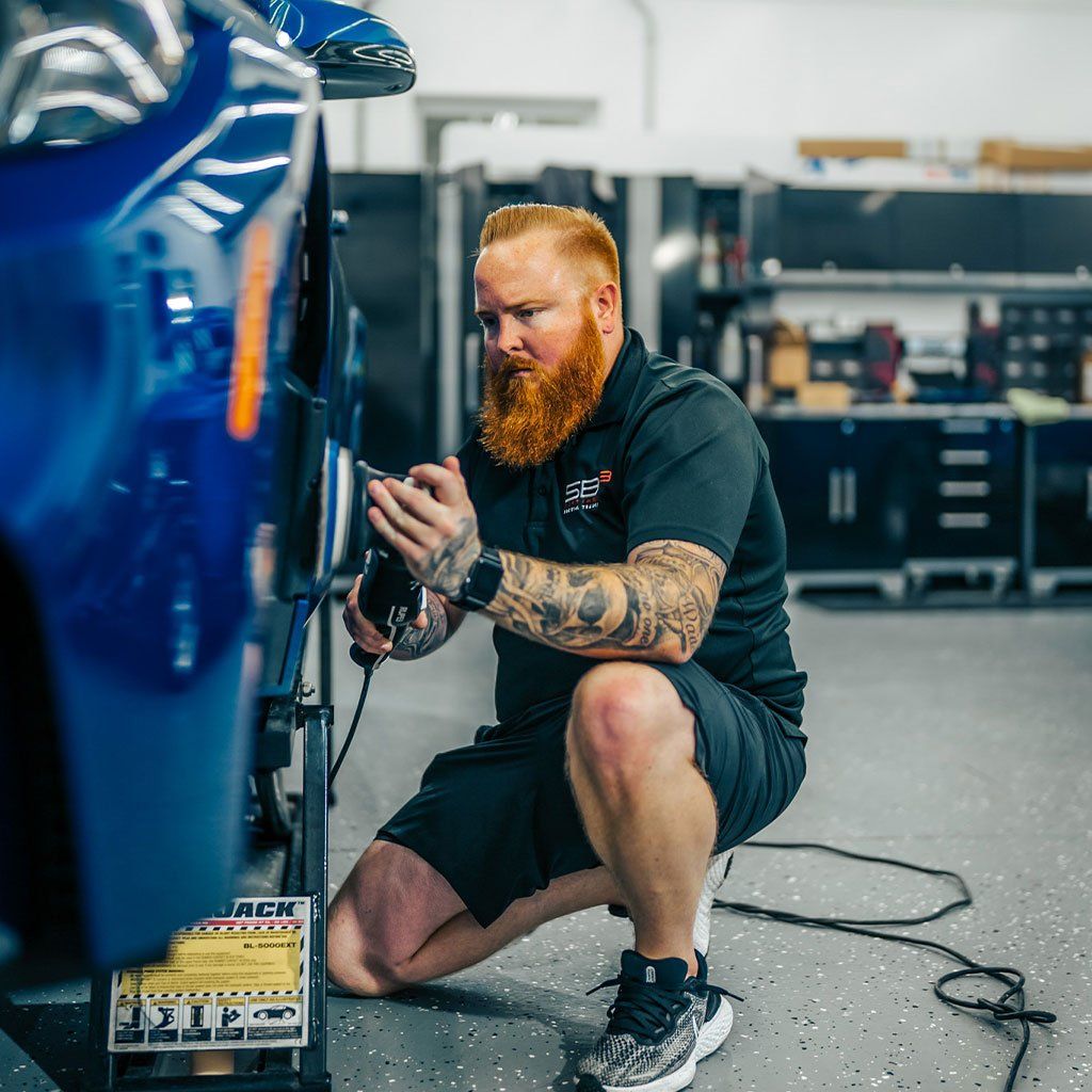 Man polishes a blue car in a shop. He kneels, using a power buffer on the car's side, with a red beard.