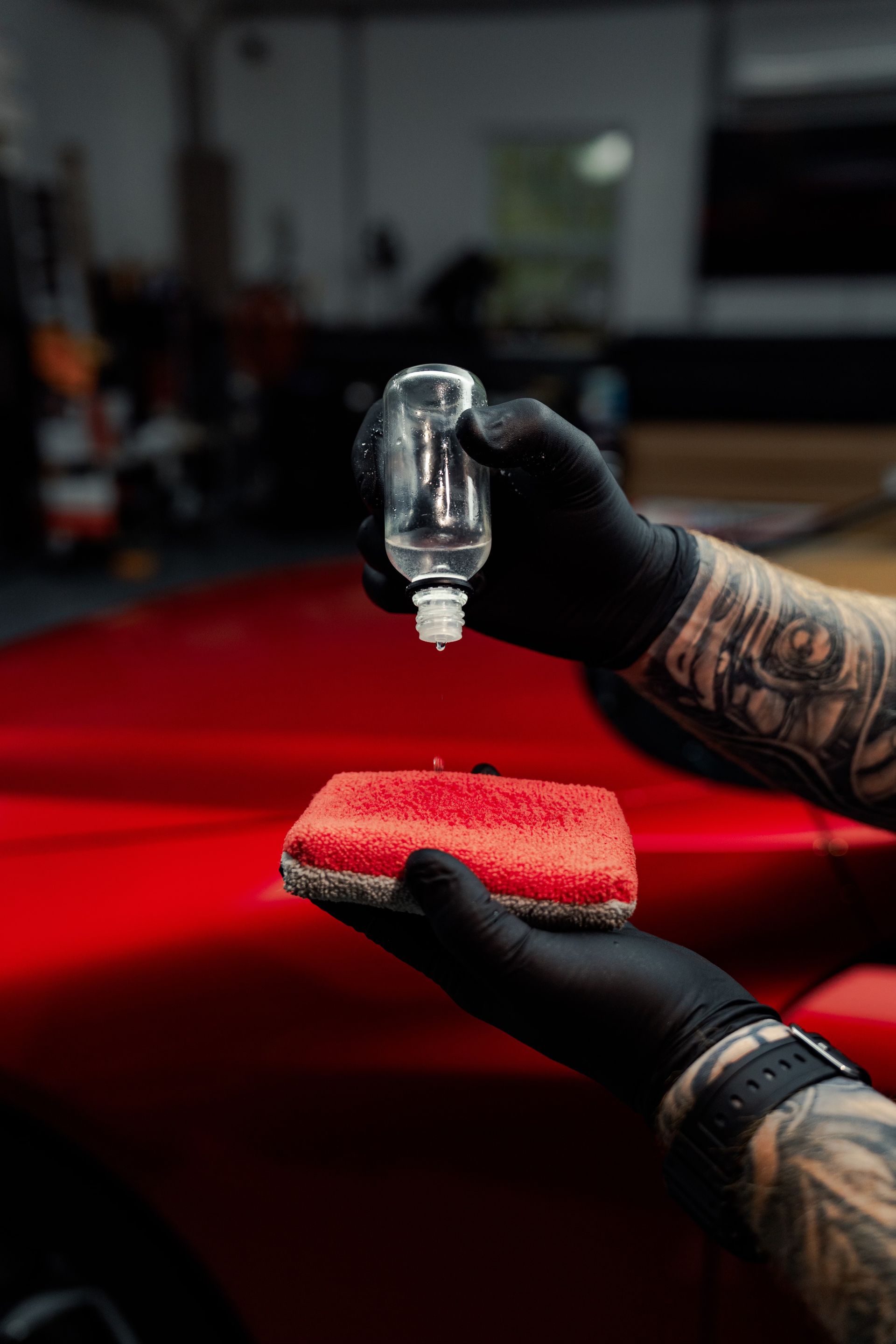 Person in black gloves dripping liquid from a bottle onto a red sponge held over a red car.