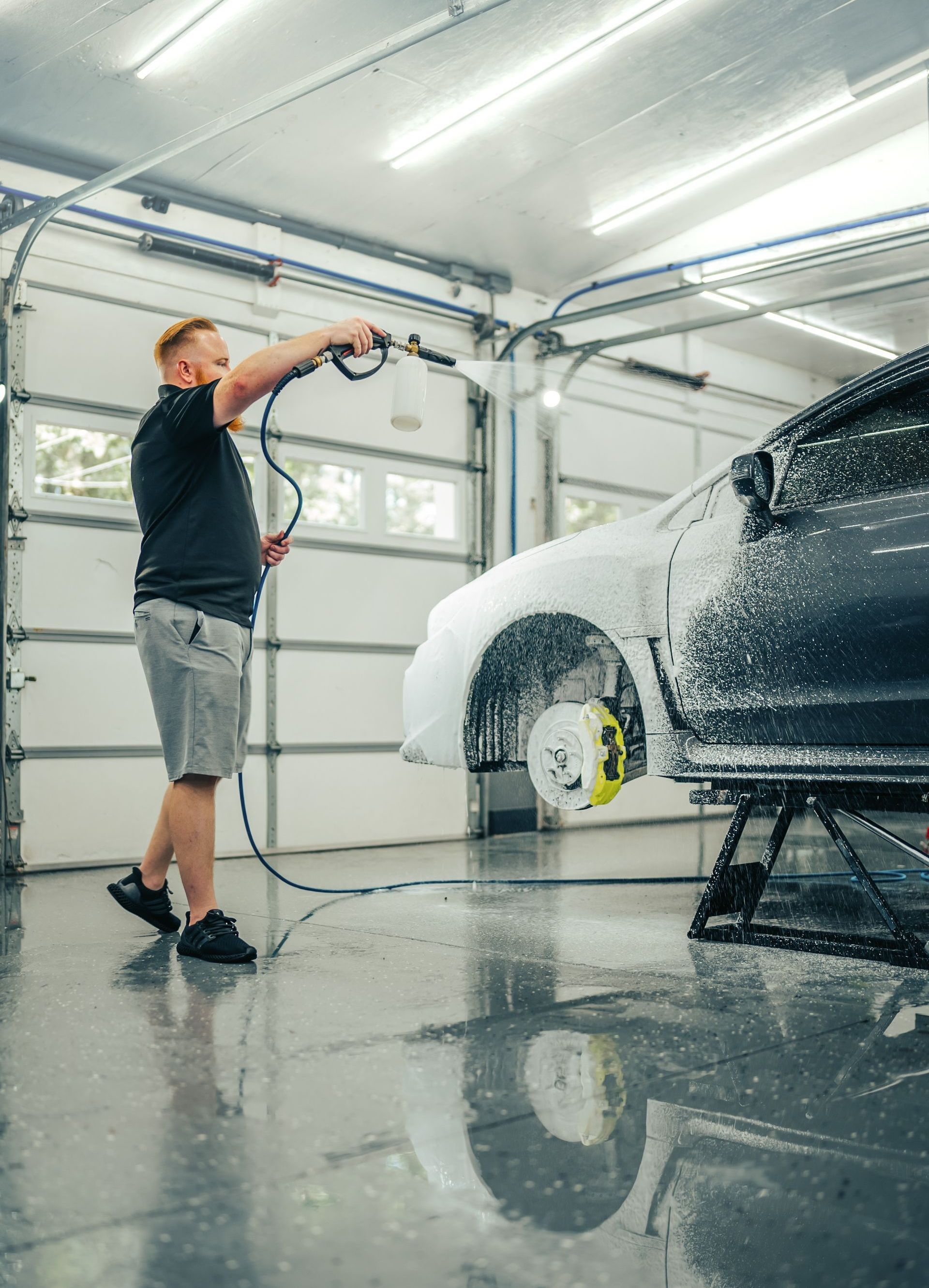 Man washing a car with foam in a garage. Car is on stands, and yellow brake calipers are visible.