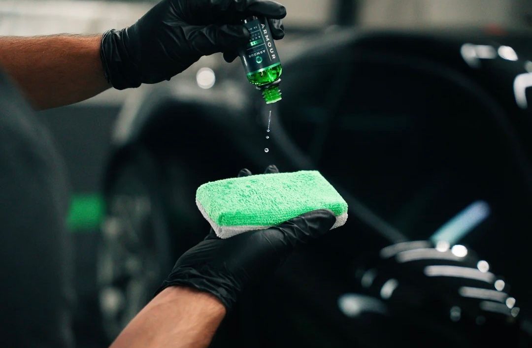 Hands in black gloves applying green liquid to a sponge on a black car.