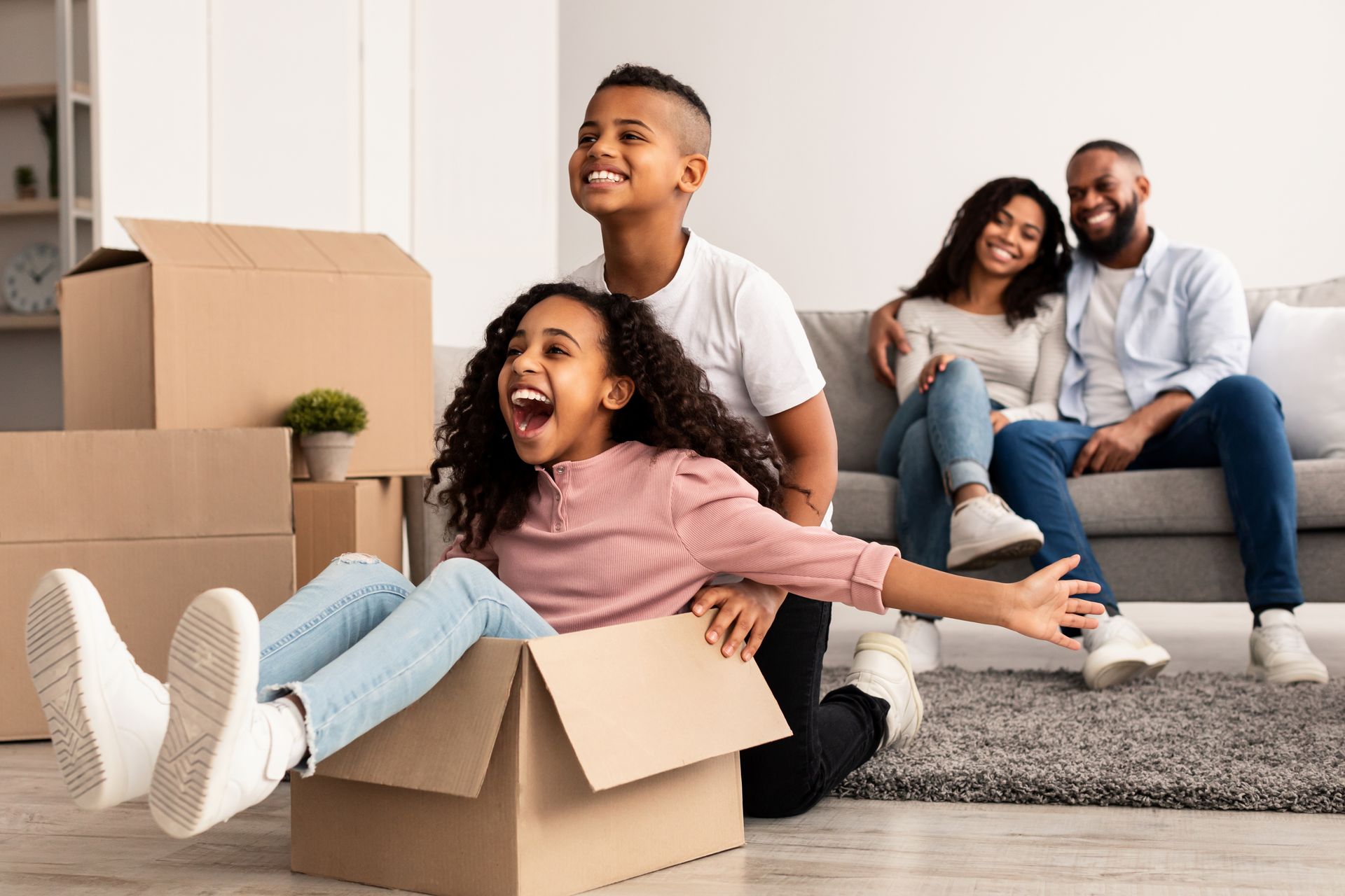 Family in new home, child in box, smiling, parents on couch, boxes in room.