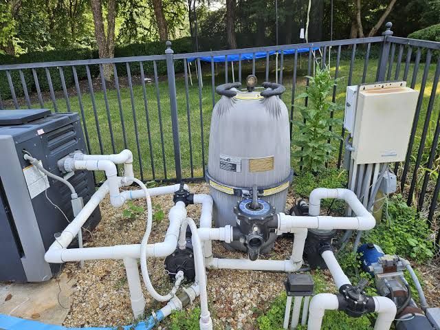 Pool filtration equipment with pipes and pump beside a fence in a wooded yard