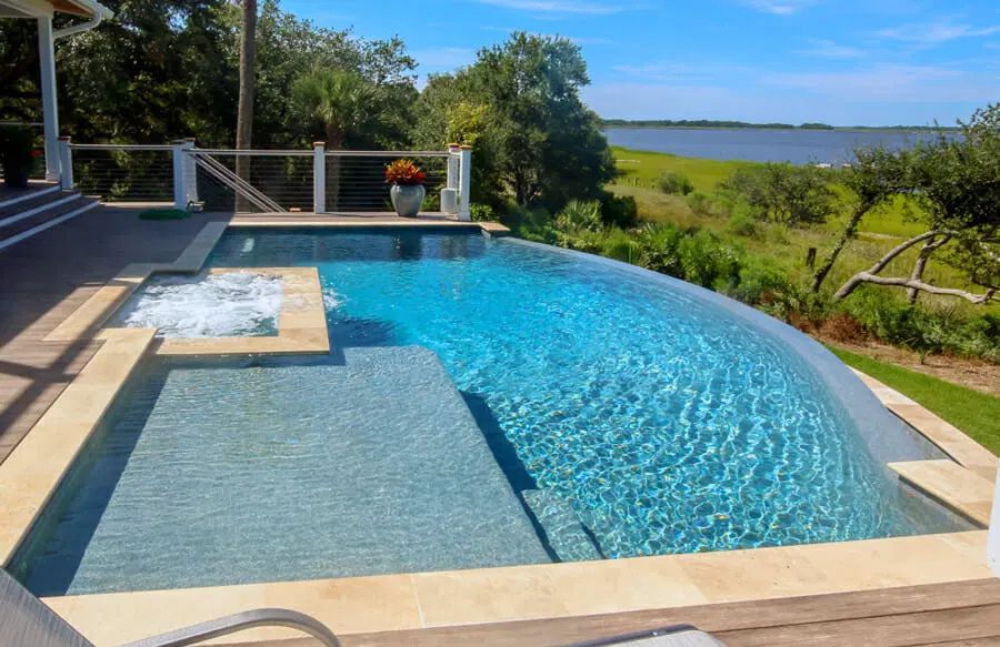 Curved blue pool with tanning ledge overlooking a lush waterfront landscape under a clear sky
