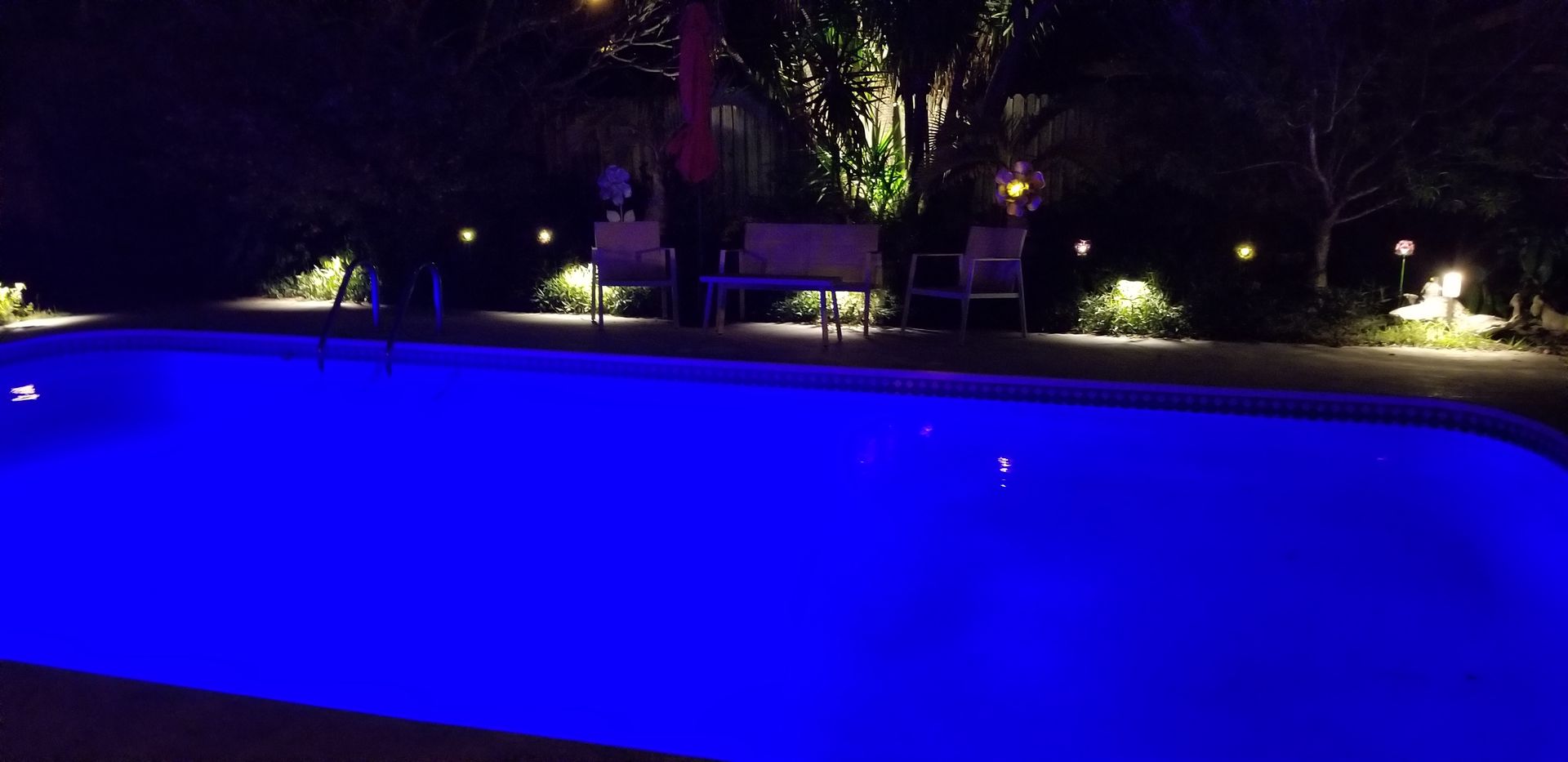 Blue-lit swimming pool at night with patio lights and trees in the background