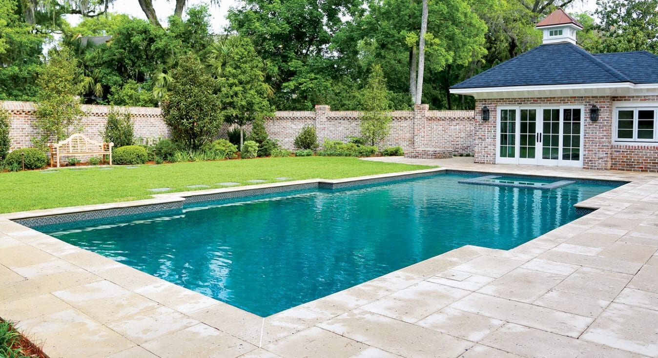 Backyard swimming pool with turquoise water beside a patio and pool house, surrounded by greenery