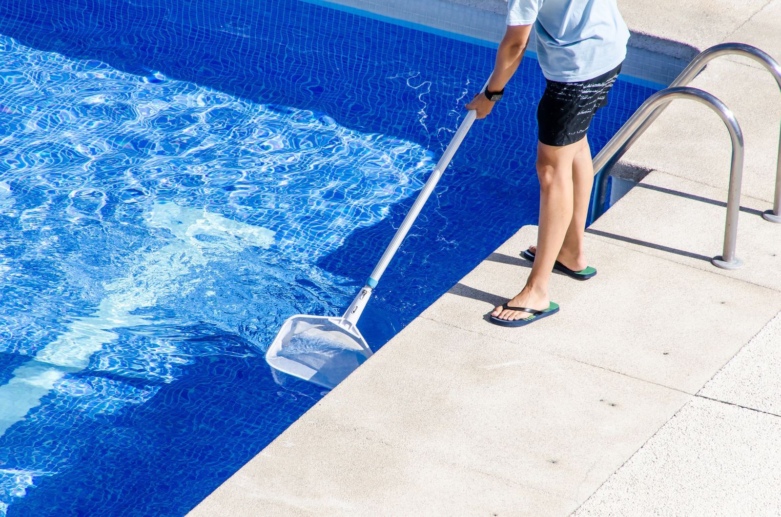Person skims a pool with a net beside blue water and a metal ladder.
