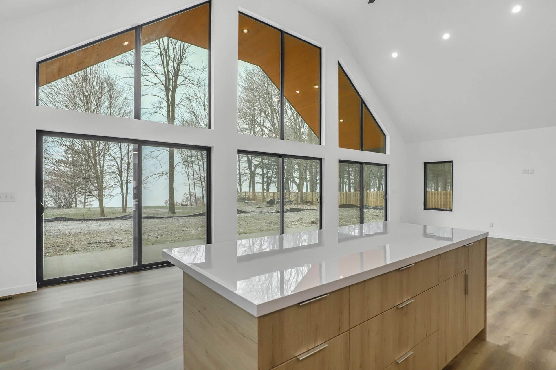 Bright modern kitchen with island, white walls, wood cabinets, and large windows/doors overlooking trees