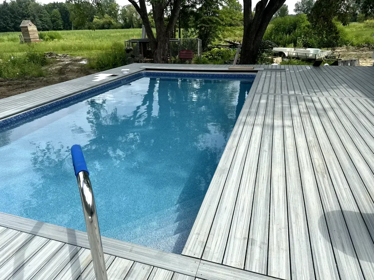 Backyard swimming pool with blue water and a wooden deck, bordered by trees and grass