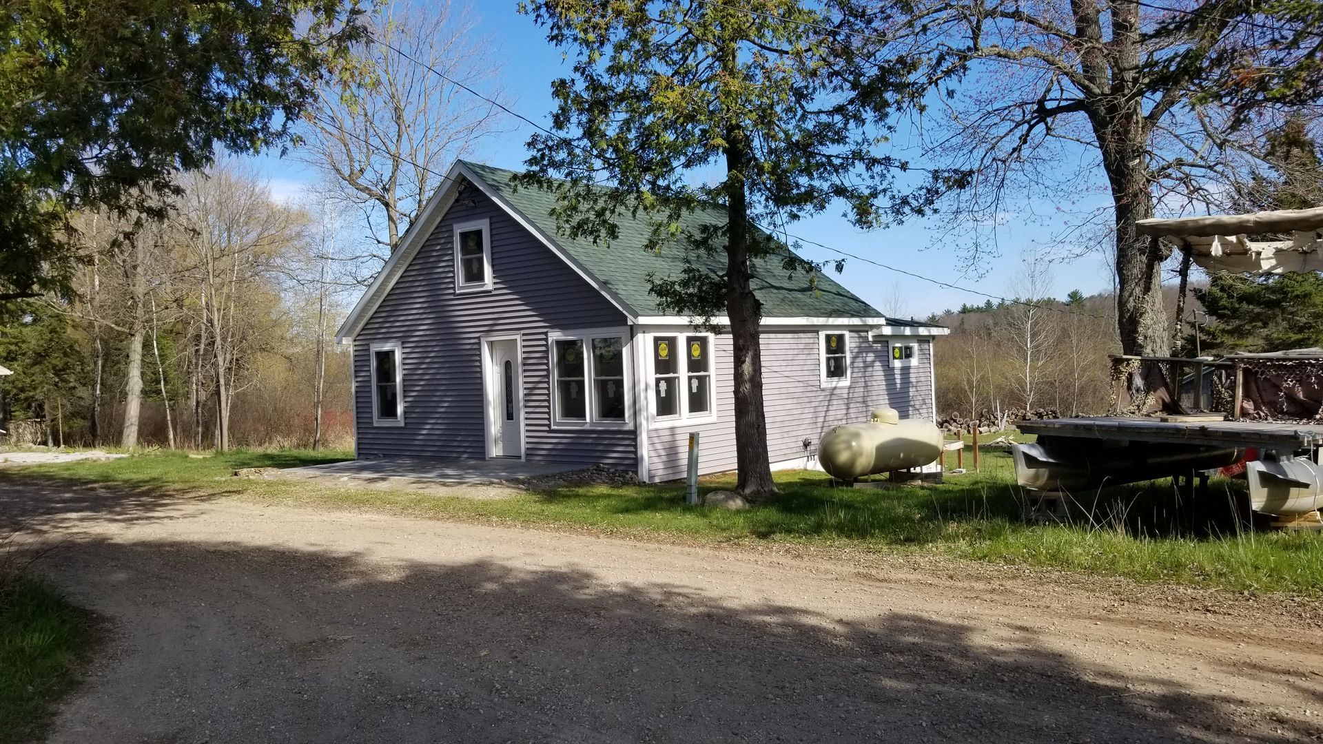 Small gray house with a porch and white trim on a gravel driveway, surrounded by trees on a sunny day