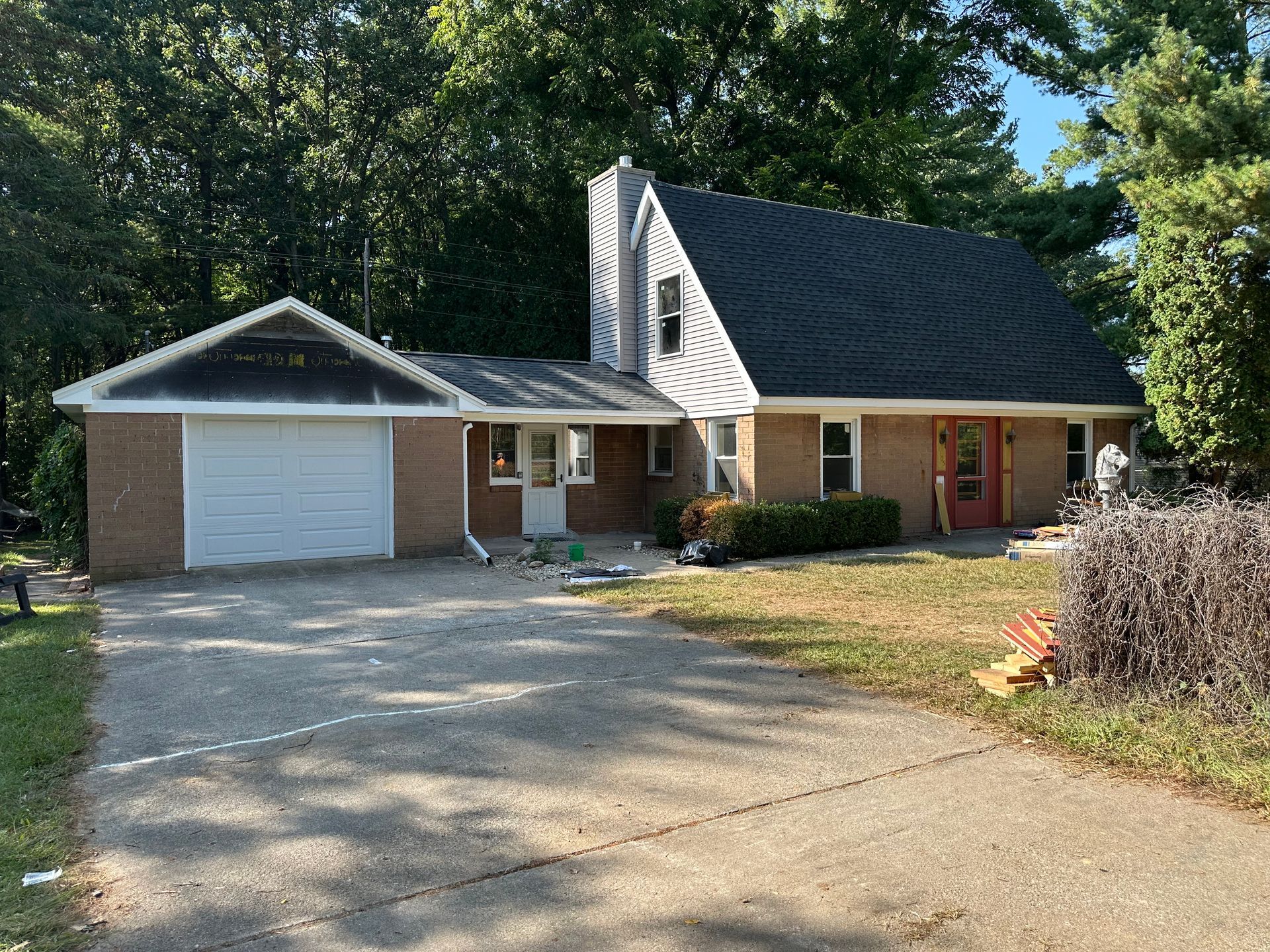 Single-story brick house with a white garage, steep dark roof, and gravel driveway surrounded by trees.