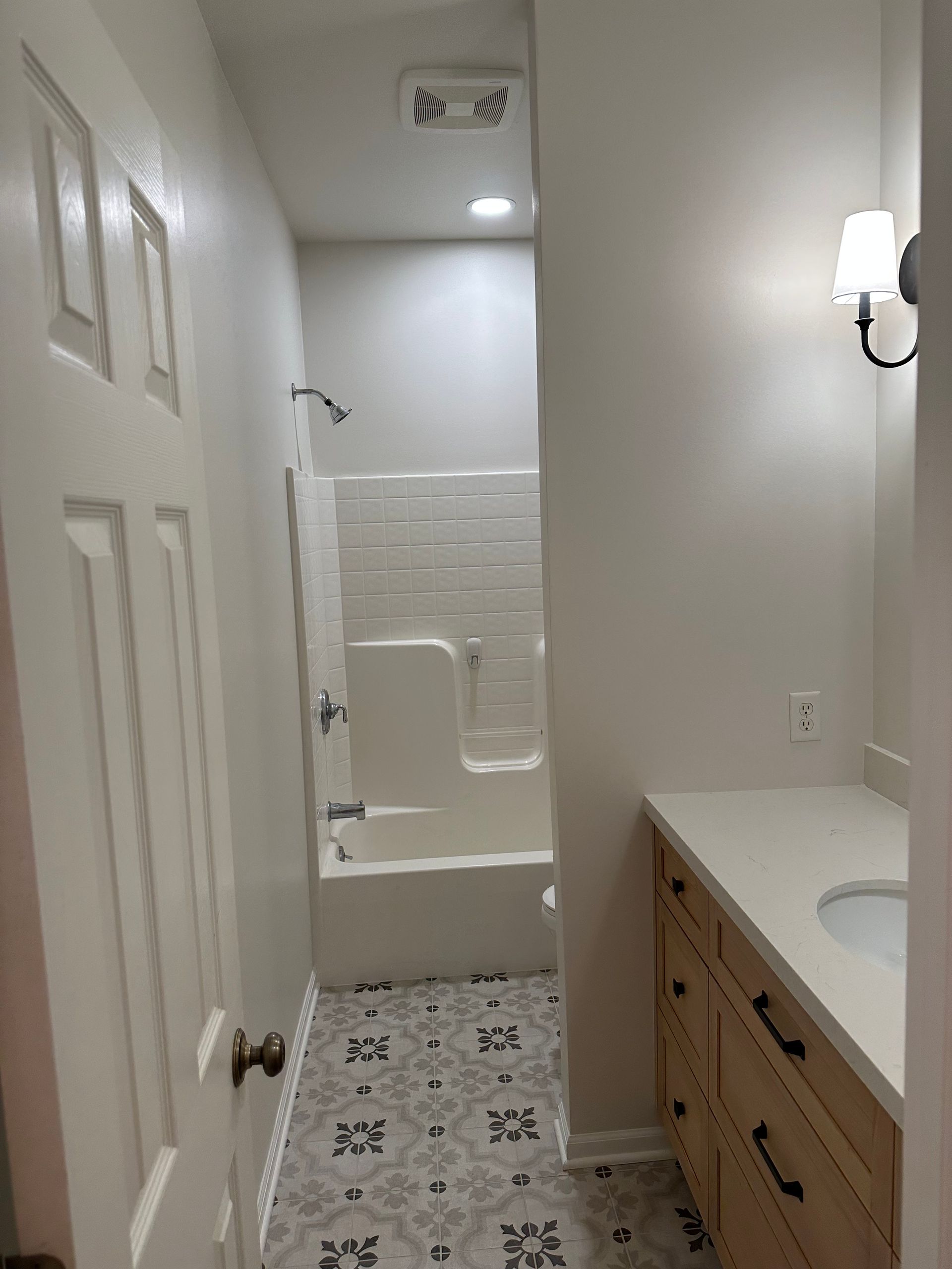 Narrow bathroom hallway with white walls, tiled floor, vanity sink, and bathtub visible at the end.