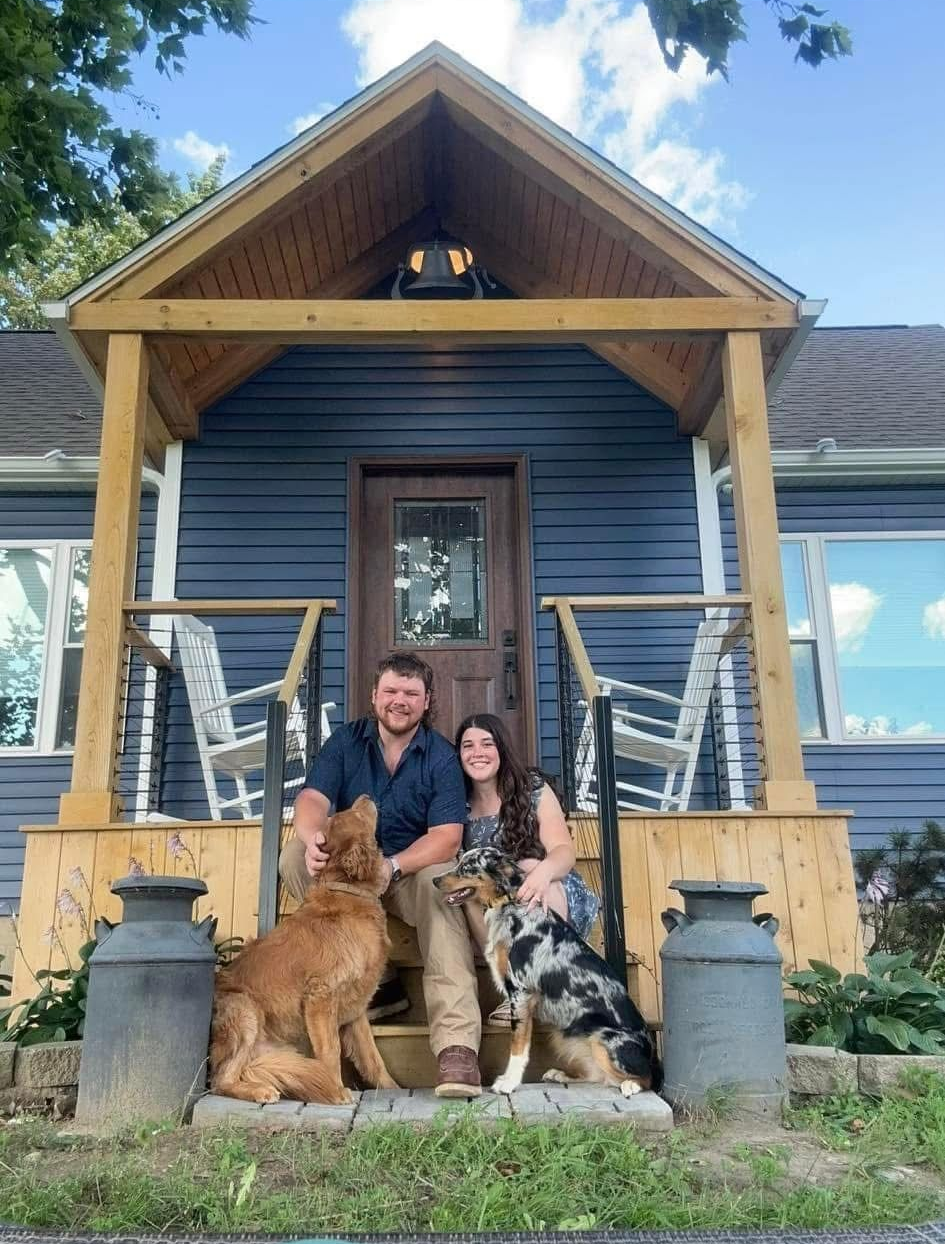 Couple seated with three dogs on a porch in front of a blue house