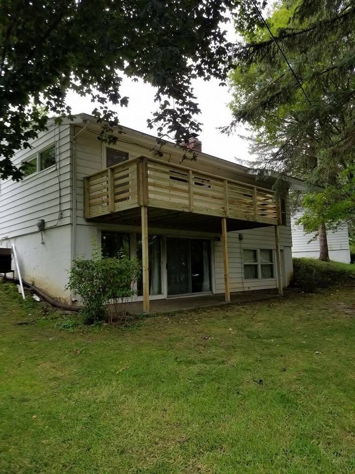 Two-story white house with a wooden balcony and porch, viewed from a grassy yard under trees