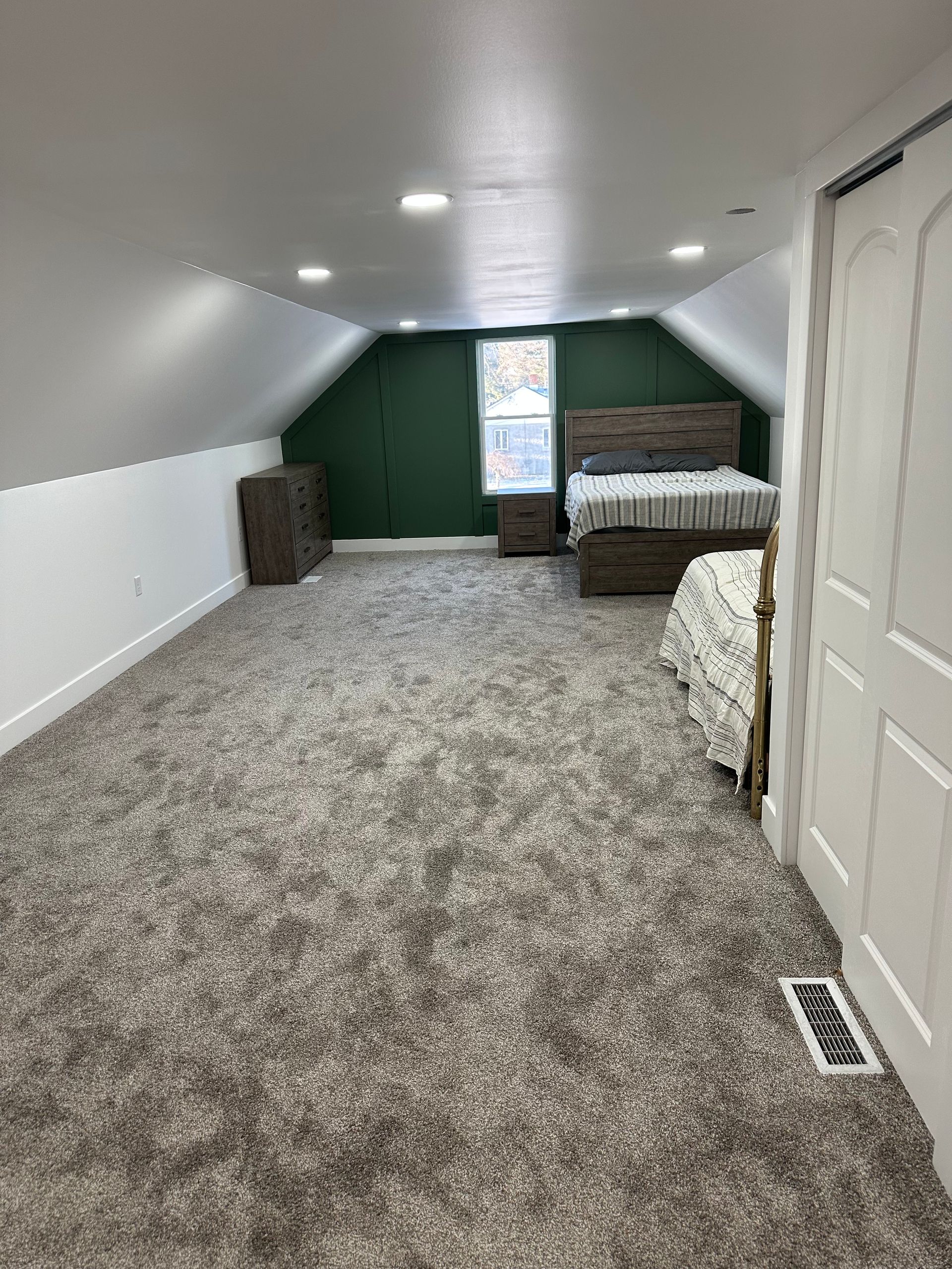 Attic bedroom with sloped ceiling, gray carpet, green accent wall, and bed by a small window.
