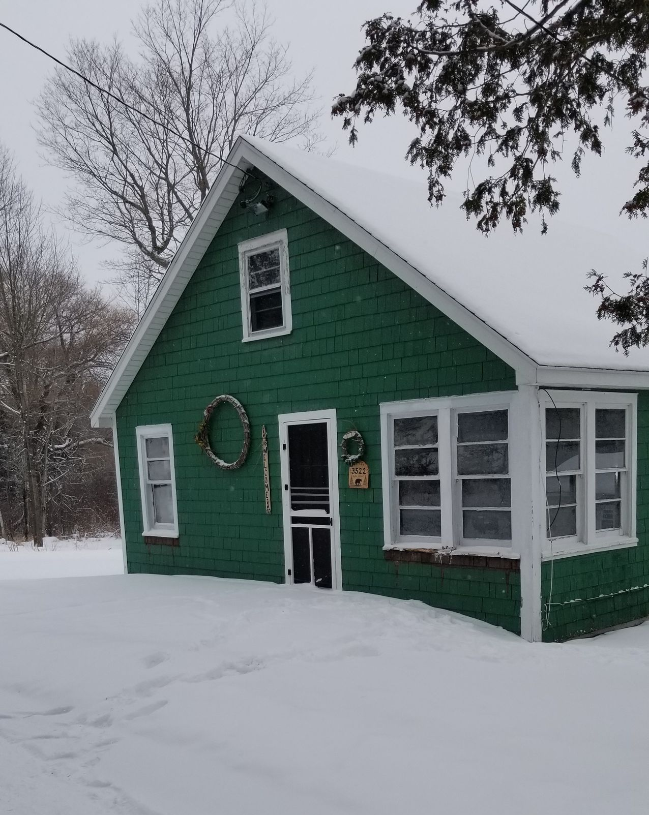 Green house with white trim in deep snow, viewed from the front, with a wreath on the door.