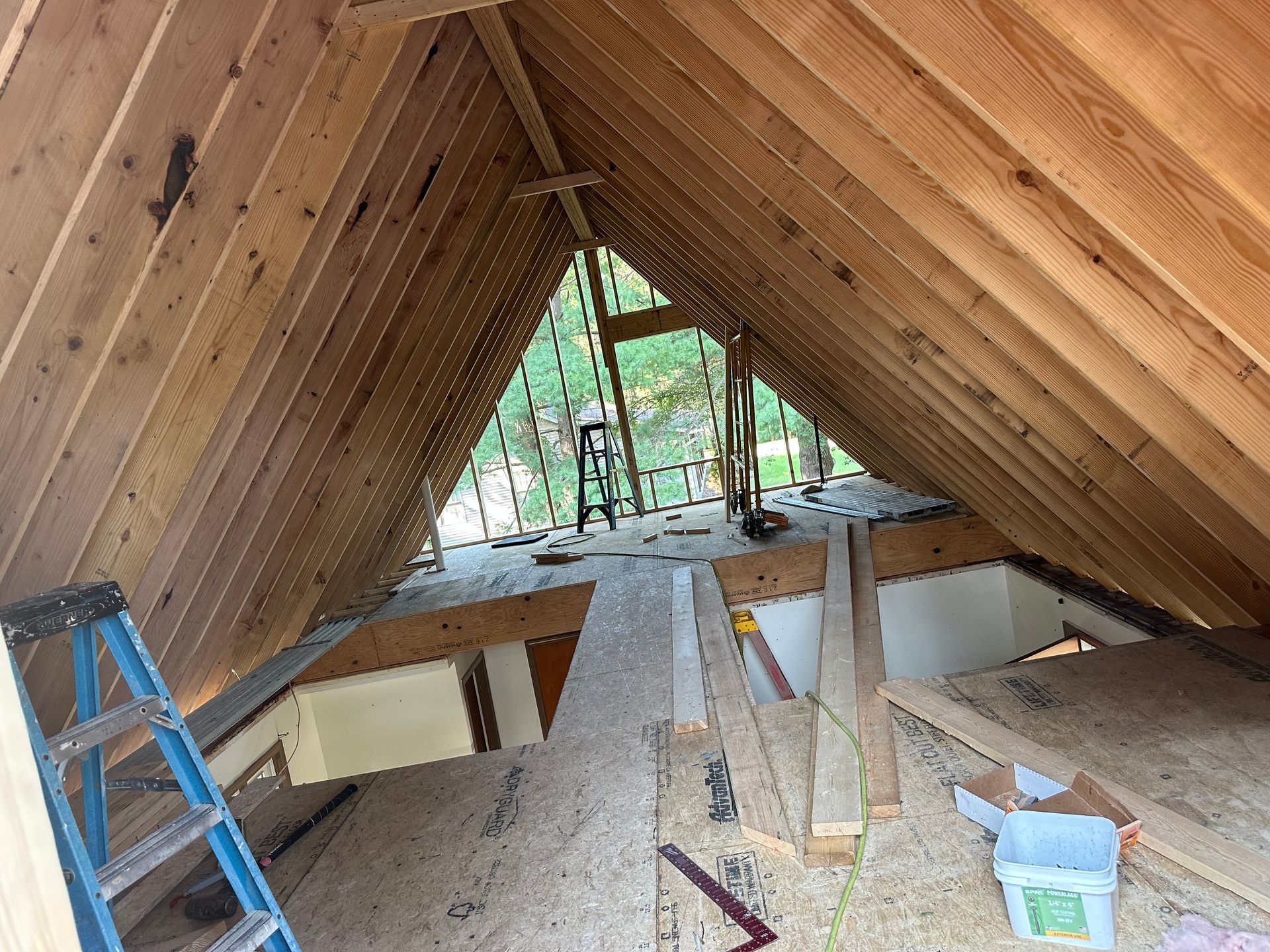 Attic under construction with wood-paneled rafters, open floor, ladder, tools, and a window view at the far end