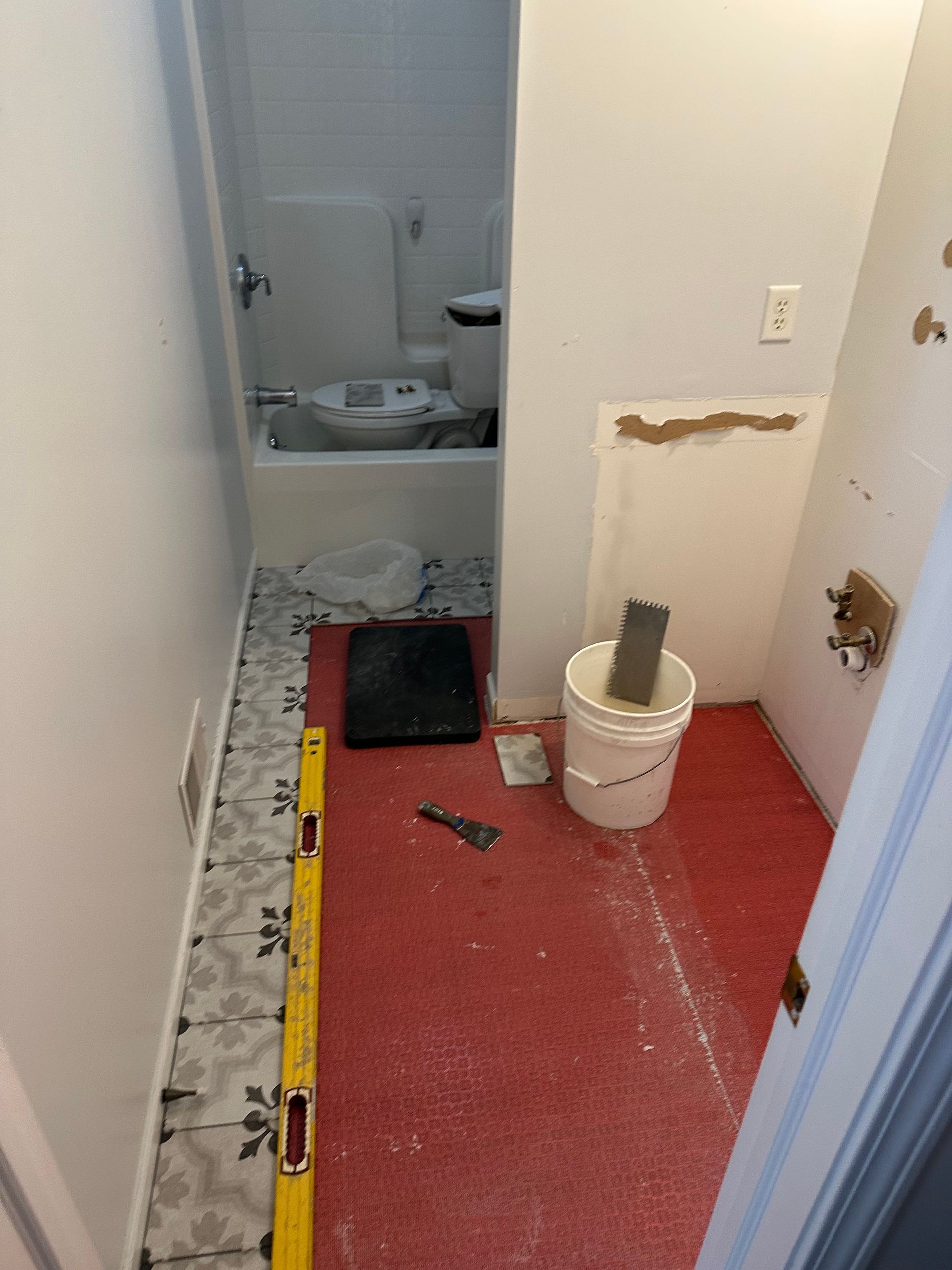 Narrow hallway under renovation with red floor covering, paint bucket, tools, and a bathroom in the back