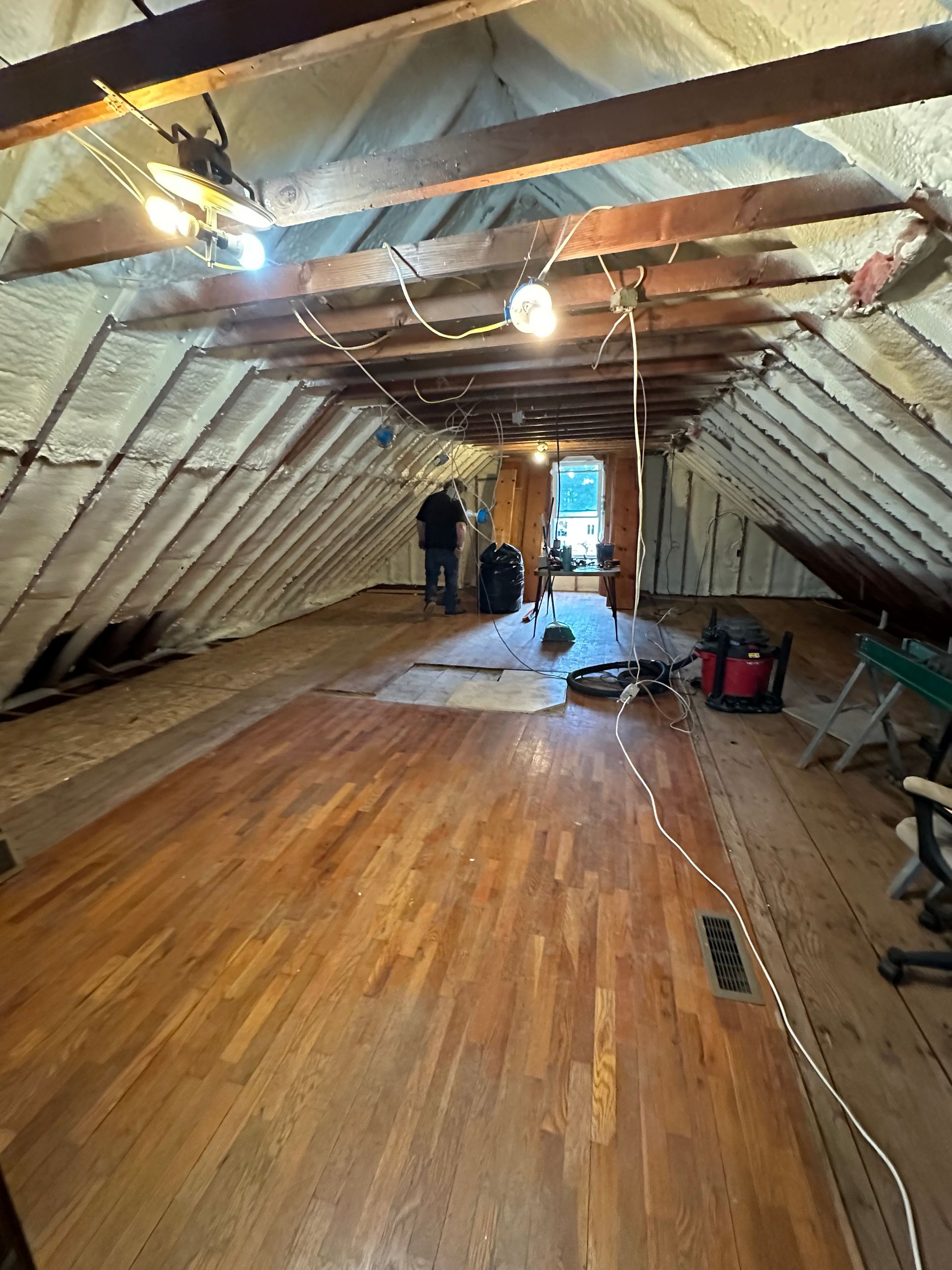 Attic renovation with exposed beams, wooden floorboards, tools, and two people working near the far end.