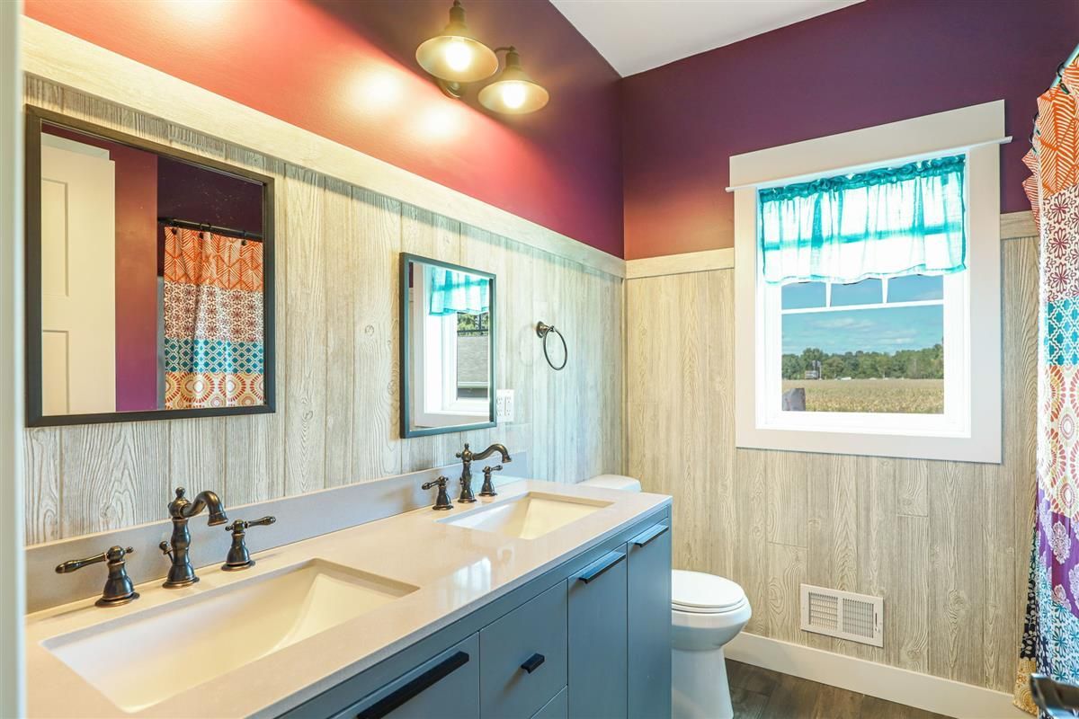 Modern bathroom with double sink, purple walls, white beadboard, and a window overlooking greenery