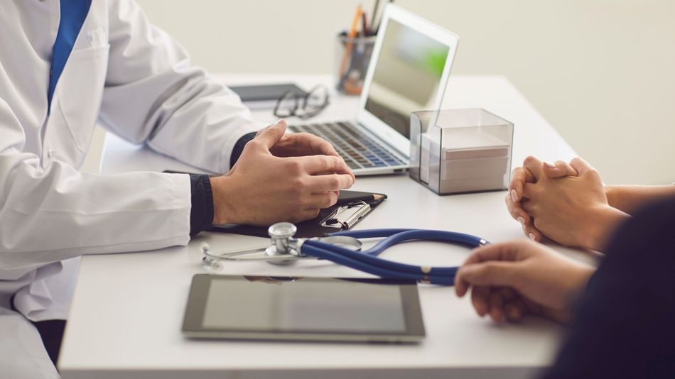 Doctor consulting with a patient at a desk, with laptop, tablet, and stethoscope visible.