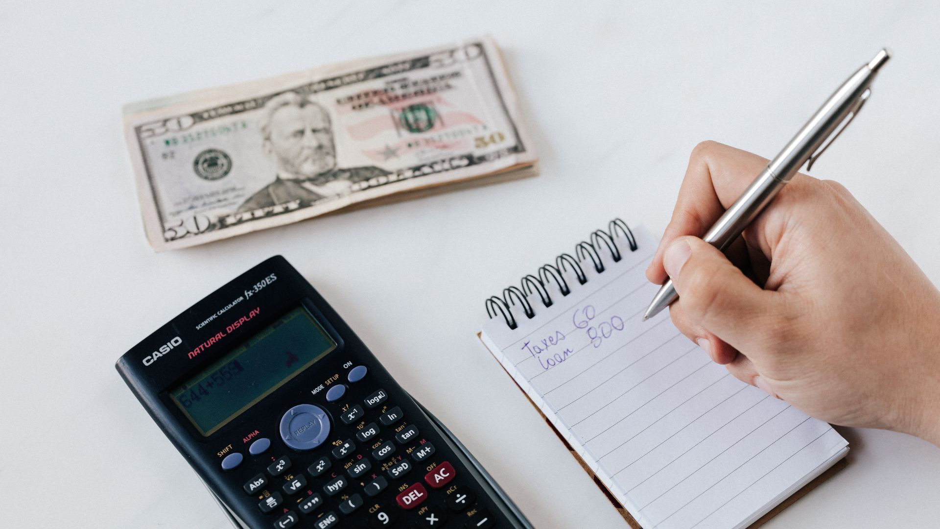 Hand holding pen over notepad beside calculator and $100 bill on a white desk
