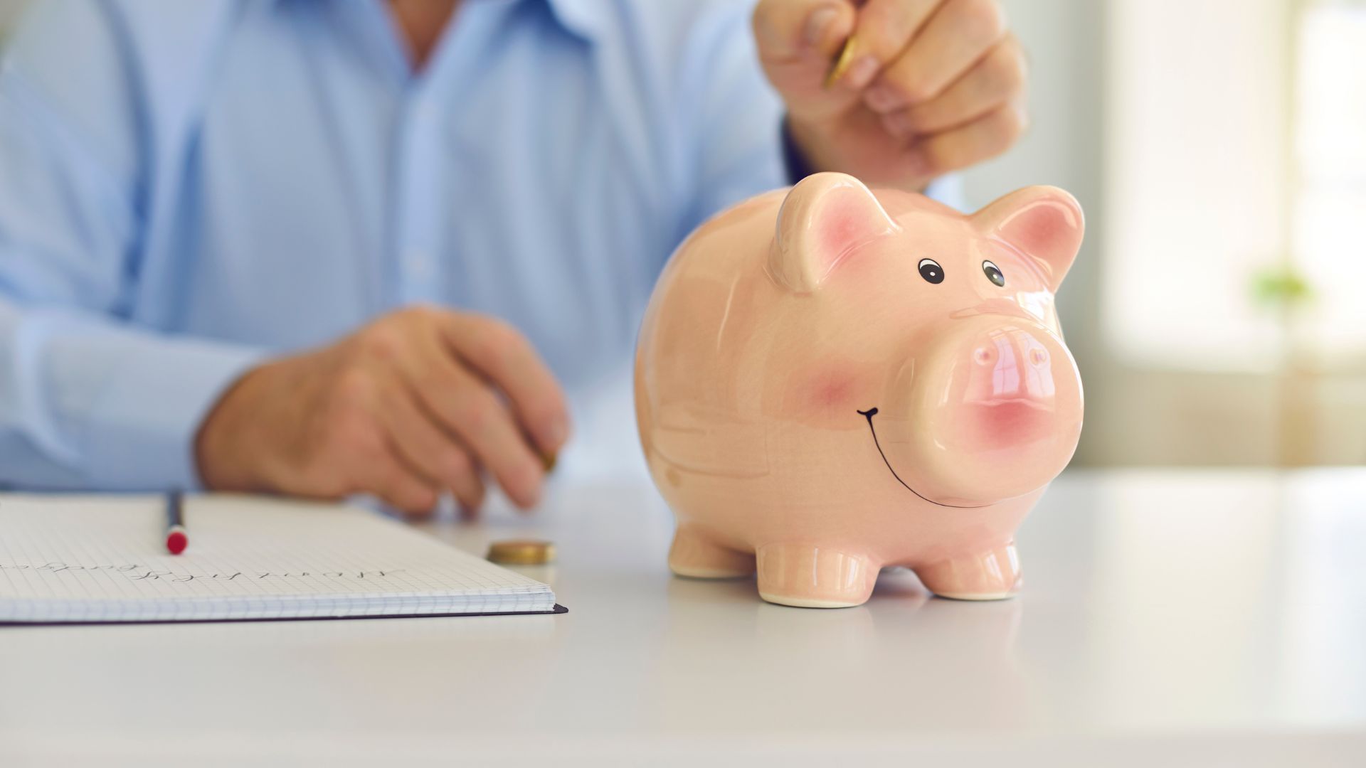 Person placing a coin into a pink piggy bank beside a notebook on a desk