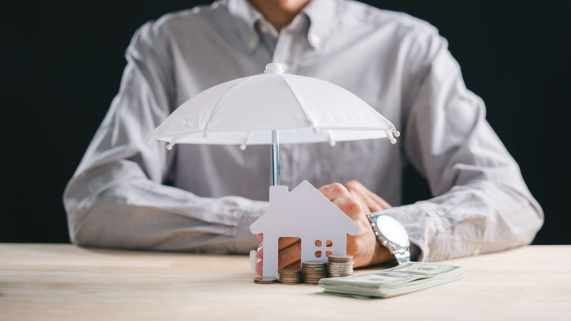 Hand placing a small house under a white umbrella beside a stack of cash on a table