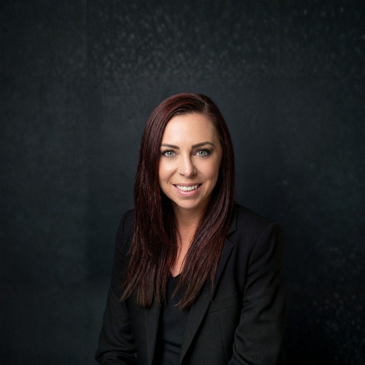 Professional headshot of a smiling person in a black blazer against a dark textured background