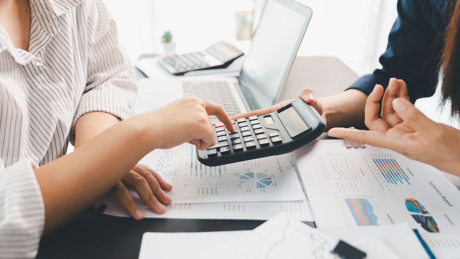 Two people use a calculator over financial documents beside a laptop in a bright office.