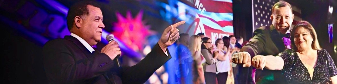 Speaker on stage gestures toward a cheering crowd with colorful lights and an American flag backdrop