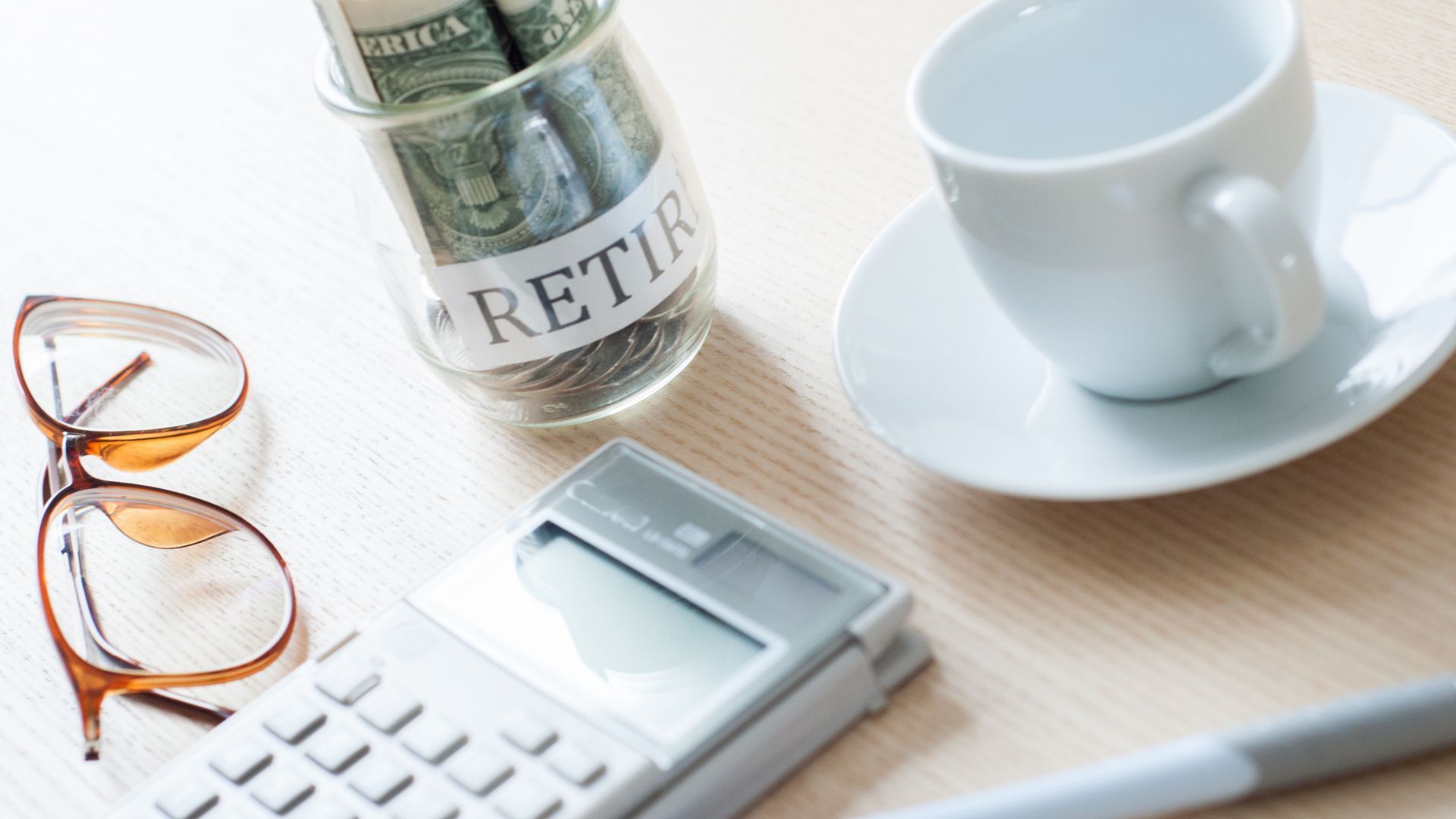 Desk with glasses, calculator, retirement jar, and coffee cup on a light wooden surface