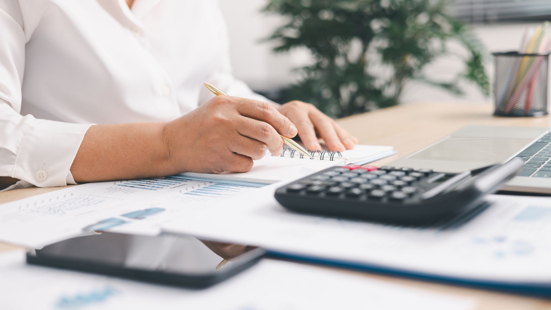Person writing on papers at a desk with a calculator and smartphone nearby