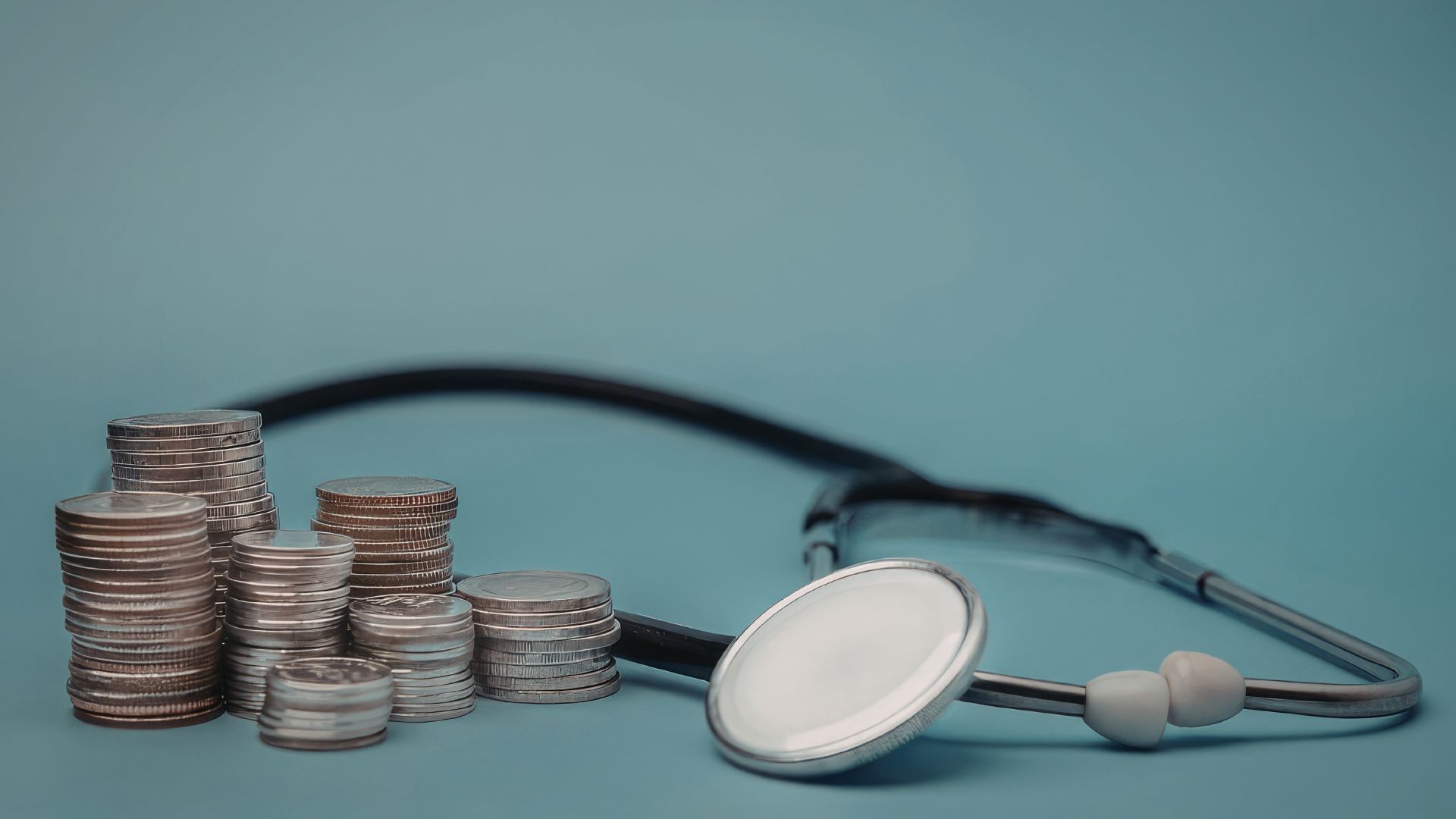 Stacks of coins beside a stethoscope on a blue background, symbolizing healthcare costs