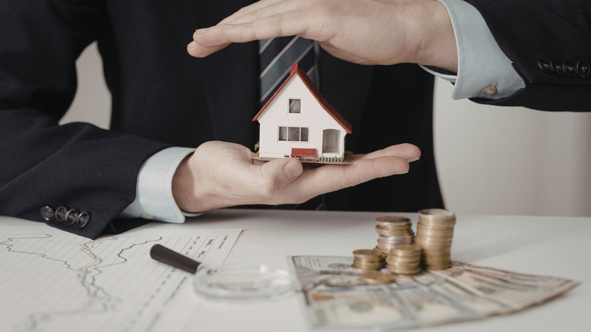 Hands holding a small house model over documents, coins, and keys on a desk.